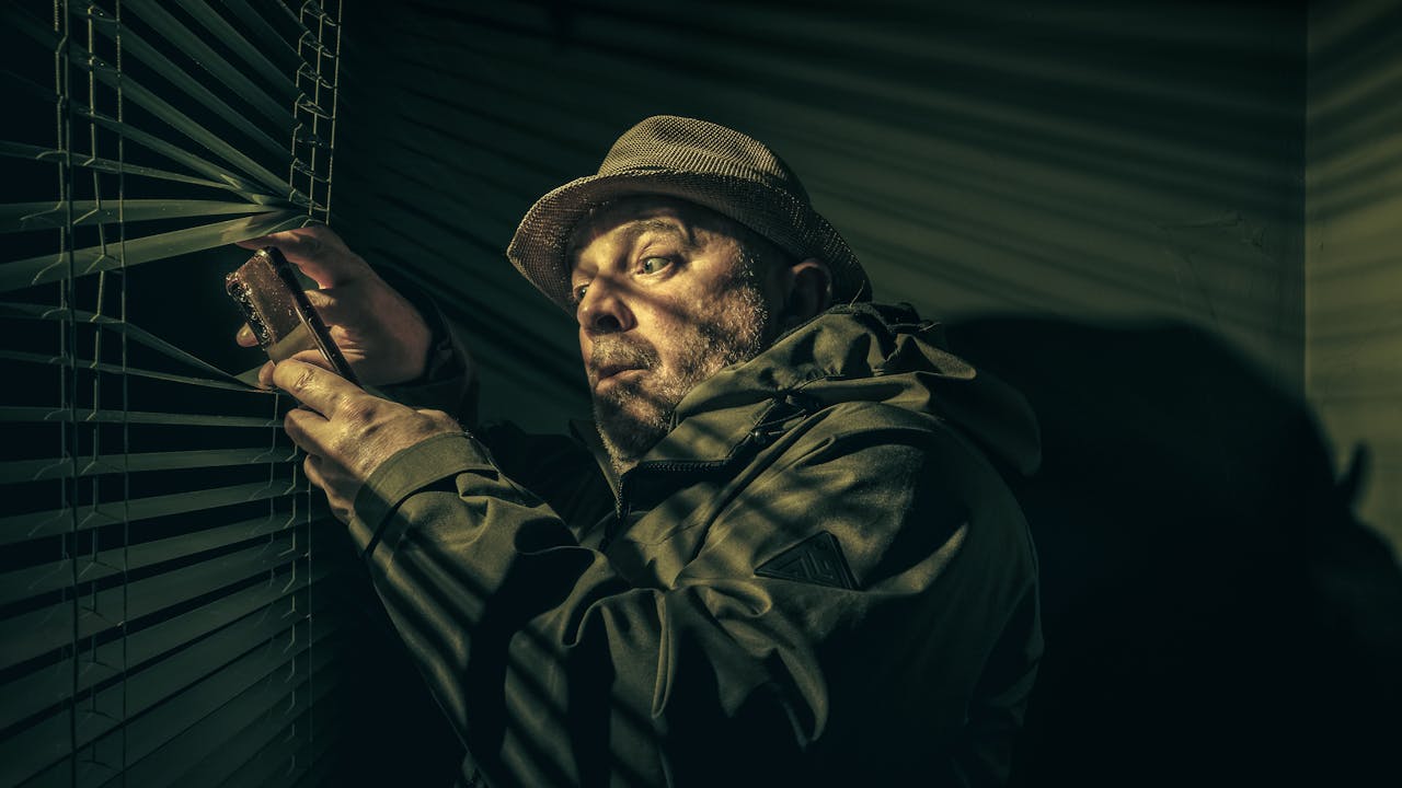 creative A man in a hat suspiciously peering through blinds, holding a phone in shadowy light.